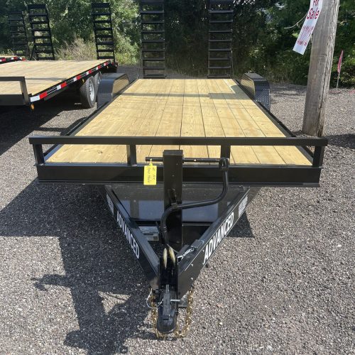 A 14k Equipment Trailer with wooden planks and a metal frame is parked on gravel by a utility pole. In the background, a Trailer for Sale sign appears through trees and shrubs, suggesting opportunities in Ohio.