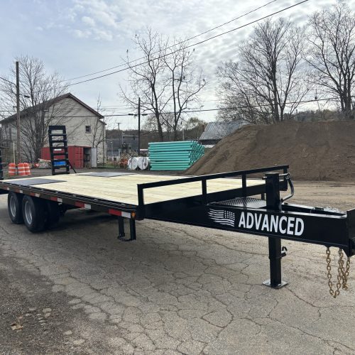 A 20k Tandem Dual Gooseneck Trailer rests on a cracked road. Behind it are stacks of pipes, a sand pile, traffic cones, bare trees, and small buildings under a cloudy sky.
