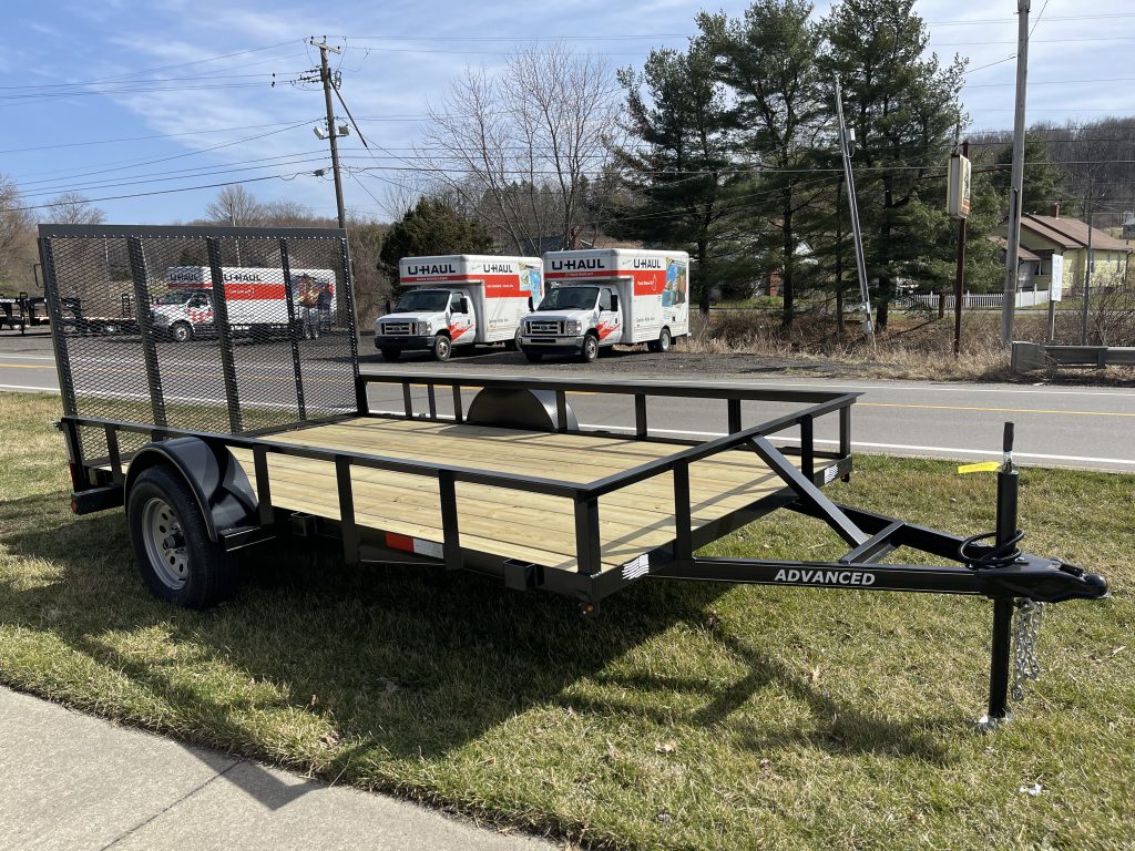 A Single Axle Utility Trailer with wooden decking and a wire mesh ramp is parked on grass near a sidewalk, with U-Haul trucks visible in the background along a tree-lined Ohio road with power lines.