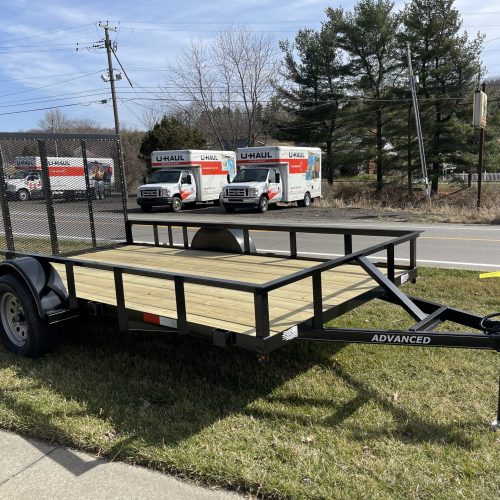 A Single Axle Utility Trailer with wooden decking and a wire mesh ramp is parked on grass near a sidewalk, with U-Haul trucks visible in the background along a tree-lined Ohio road with power lines.