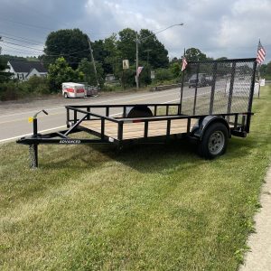A Single Axle Utility Trailer is for sale, parked on grass beside a road in Ohio. It has a black metal frame, wooden boards, a mesh ramp, and small American flags at the back. A residential area and cloudy sky serve as the backdrop.
