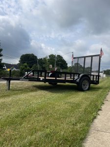 A Single Axle Utility Trailer with a metal grid ramp and two American flags is parked on a grassy area near an Ohio sidewalk. Under the cloudy sky, trees and utility poles complete the scene as this trailer for sale awaits its next adventure.