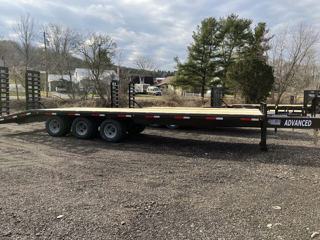 A large flatbed trailer with multiple wheels, parked on a gravel area in Ohio, catches the eye. This black trailer, labeled ADVANCED, is available for sale. Trees and a few buildings peek from the background under a cloudy sky.