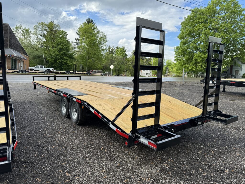 A flatbed trailer with a wooden deck and dual axles is parked on a gravel surface, ready for sale in Ohio. In the background, lush green trees, a small building, and a cloudy sky complete the scene. The trailer features two loading ramps at the rear, perfect for versatile hauling needs.