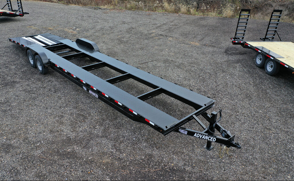 An empty, long flatbed trailer with a black metal frame is parked on a gravel surface in Ohio. It features red and white reflective markings on the sides and is labeled Advanced at the hitch. Trailer for sale signs are visible while other trailers appear partially in the background.