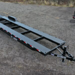 An empty, long flatbed trailer with a black metal frame is parked on a gravel surface in Ohio. It features red and white reflective markings on the sides and is labeled Advanced at the hitch. Trailer for sale signs are visible while other trailers appear partially in the background.