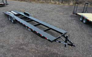 An empty, long flatbed trailer with a black metal frame is parked on a gravel surface in Ohio. It features red and white reflective markings on the sides and is labeled Advanced at the hitch. Trailer for sale signs are visible while other trailers appear partially in the background.