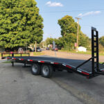 A dual axle flatbed trailer, perfect for those in Ohio seeking reliability, is parked on the street. This trailer for sale boasts a black metal frame and a rear ramp. Picturesque trees and houses stand in the background under a clear blue sky.