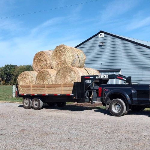 A black pickup truck is hitched to a 14K Gooseneck Deckover Dump Trailer loaded with large round hay bales. The trailer is parked on a gravel area in front of a metal barn, with a grassy field and clear blue sky in the background.