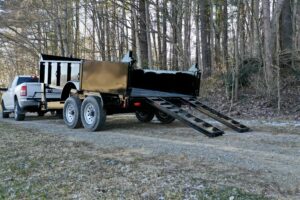 A pickup truck tows a black 14K Gooseneck Low Profile Dump Trailer with its rear gate open and metal loading ramps extended on a dirt and gravel path by a forested area.