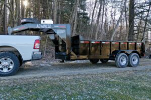 A white pickup truck tows a black 14K Gooseneck Low Profile Dump Trailer on a gravel path beside leafless trees.
