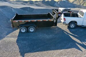 A white pickup truck is towing a 14K Gooseneck Low Profile Dump Trailer with dual axles, parked on a gravel lot surrounded by gravel piles.