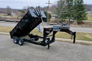 A 14K Gooseneck Low Profile Dump Trailer with dual axles, raised hydraulic bed, and spare tire mounted on the front sits on a paved surface near a road, displaying ADVANCED branding. Trees and houses are visible in the background.