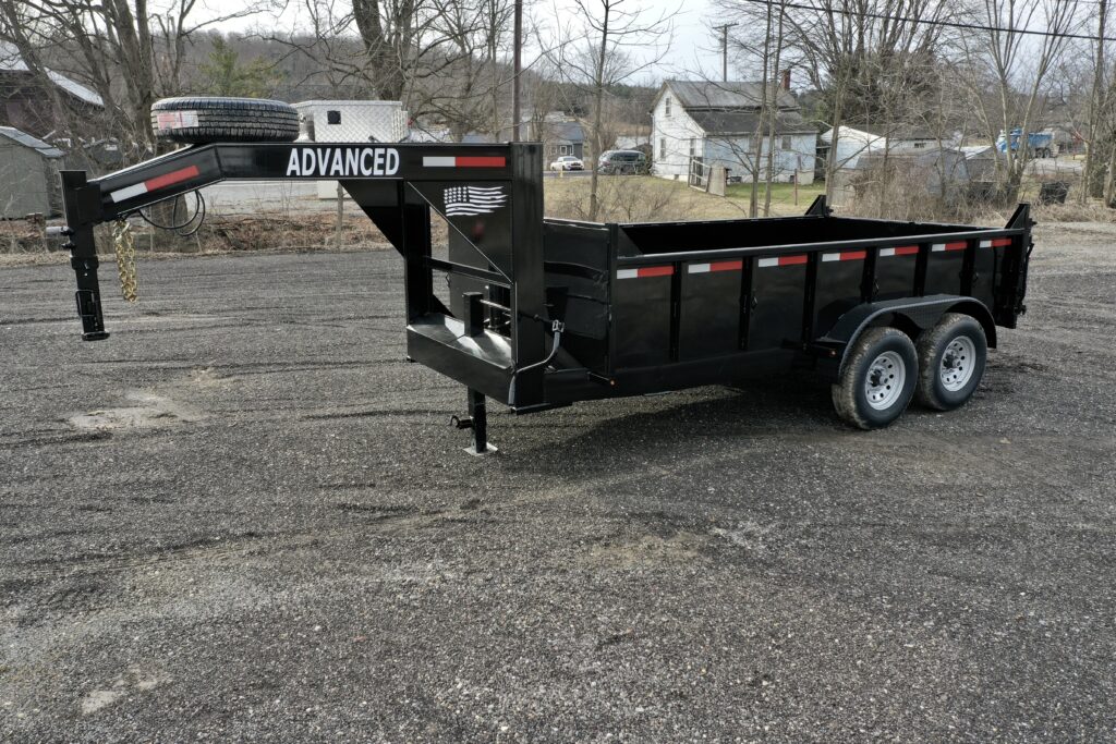 A black 14K Gooseneck Low Profile Dump Trailer with ADVANCED on the gooseneck is parked on gravel. It has dual axles, red and white reflective stripes, a front-mounted spare tire, and houses with trees in the background.