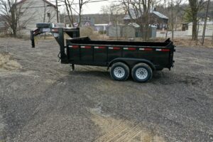 A 14K Gooseneck Low Profile Dump Trailer with dual axles is parked on a gravel lot, with trees and several buildings visible in the background on an overcast day.