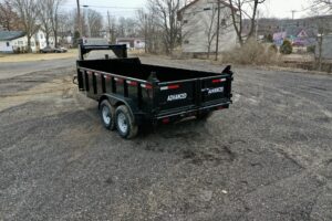 A 14K Gooseneck Low Profile Dump Trailer with dual axles is parked on a gravel lot. The trailers gate reads ADVANCED and its surrounded by houses, trees, and utility poles on a cloudy day.