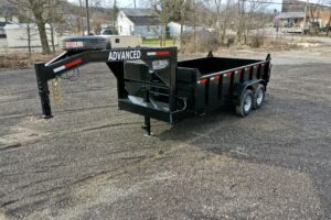 A 14K Gooseneck Low Profile Dump Trailer, black with dual axles and ADVANCED on the gooseneck hitch, sits on a gravel lot. It features red and white reflective stripes, a mounted spare tire, bare trees, and buildings in the background.