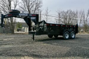 A 14K Gooseneck Low Profile Dump Trailer with dual axles is parked on gravel. It has a black, heavy-duty build and features the ADVANCED label on the front, with trees and buildings visible in the background.