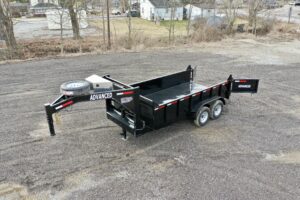 A 14K Gooseneck Low Profile Dump Trailer with dual axles and ADVANCED branding is parked on a gravel lot, with trees and houses in the background.