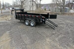A 14K Gooseneck Low Profile Dump Trailer with dual axles is parked on gravel, its rear gate open and two metal ramps extended to the ground. Houses, trees, and another trailer are visible in the background.