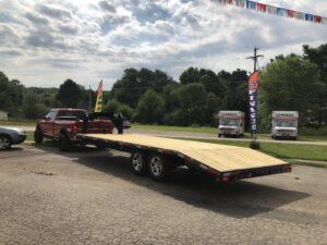 A red truck with a flatbed trailer is parked on a paved lot in Ohio under a cloudy sky. Nearby, two U-Haul trucks are stationed. Flags and trees lend color to the scene, enhancing the subtle charm of this trailer for sale setting.