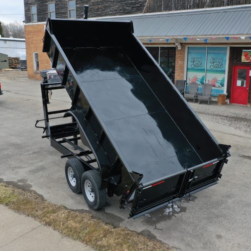 A 14K Gooseneck Low Profile Dump Trailer with its bed raised and dual wheels is parked on the asphalt street near a grass edge, in front of a brick building displaying For Sale signs in the window.