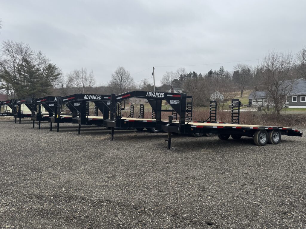 A row of three flatbed trailers, each with ADVANCED on the goosenecks and marked as trailers for sale, are parked on a gravel lot. In the background, leafless trees and a few houses sit under a cloudy sky.