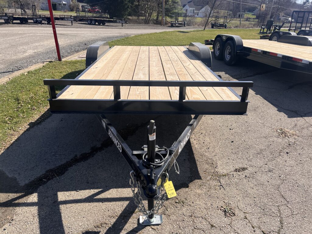 A flatbed trailer with a wooden deck is parked on a paved surface. It has a metal frame and hitch, and there are other trailers visible in the background. Some grass and a road are nearby.