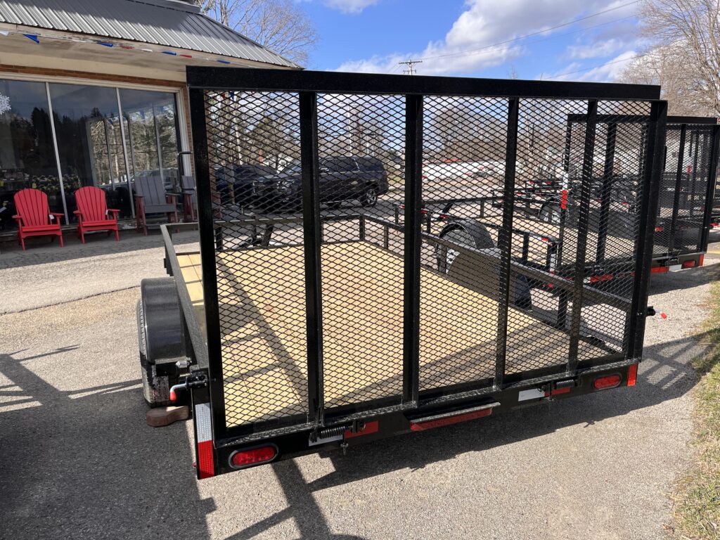 An In Stock - 12 Single Axle Utility Trailer with a black metal mesh is parked on a driveway before a building. Red chairs are seen on the left porch, under partly cloudy skies, with trees and another vehicle in the background.