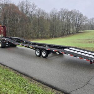 A 16k Low Pro Wedge, a black car hauler trailer, is being towed by a red semi-truck on a wet rural road with grass and leafless trees in the background under an overcast sky.