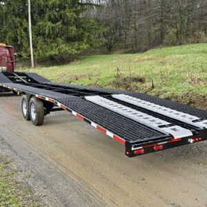 A 21k Low Pro Wedge, an empty black car hauler trailer with ramps extended, is parked on a gravel road beside a grassy, wooded area and hitched to a red semi-truck.