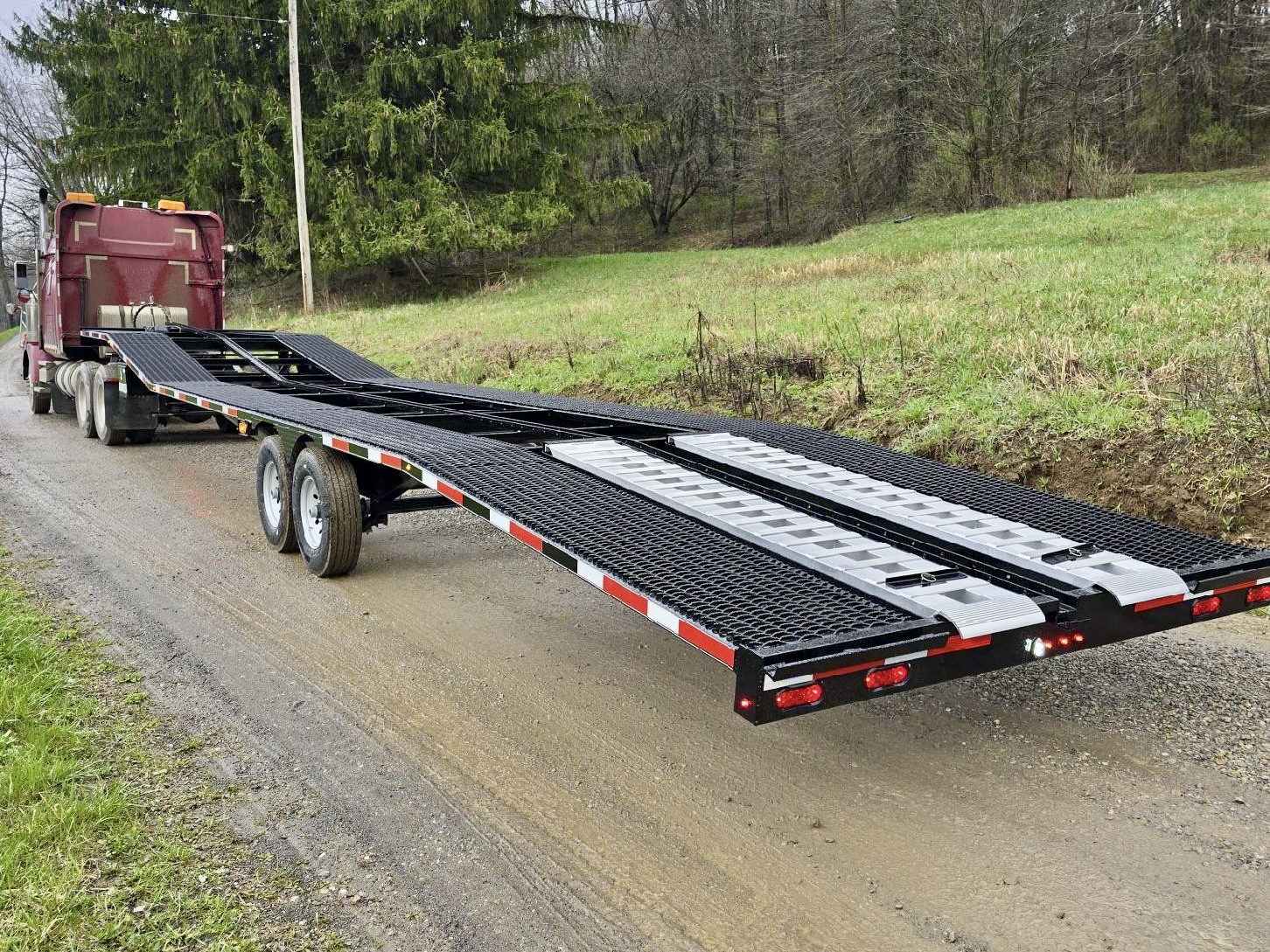 A 21k Low Pro Wedge, an empty black car hauler trailer with ramps extended, is parked on a gravel road beside a grassy, wooded area and hitched to a red semi-truck.