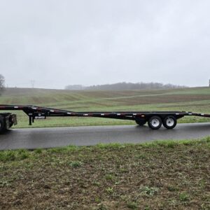 A 14k Low Pro Wedge gooseneck flatbed trailer is attached to a truck and parked on a rural road, with fields and trees in the background under an overcast sky.
