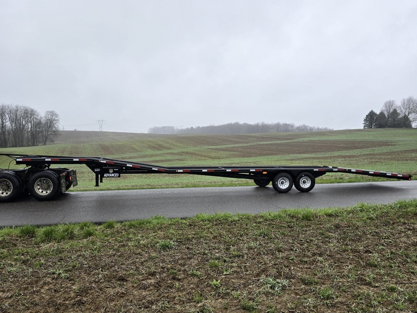 A 14k Low Pro Wedge gooseneck flatbed trailer is attached to a truck and parked on a rural road, with fields and trees in the background under an overcast sky.