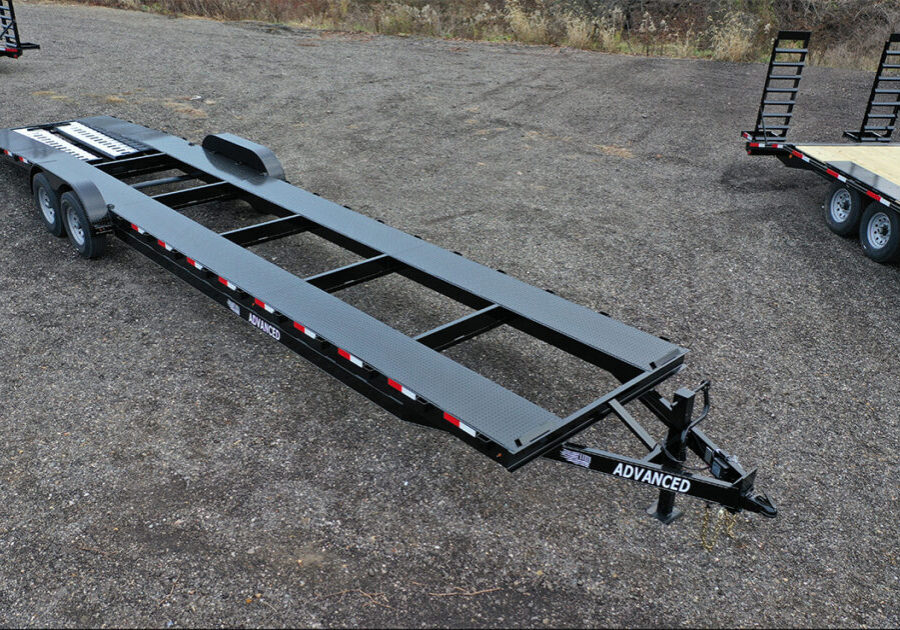 An empty, long flatbed trailer with a black metal frame is parked on a gravel surface in Ohio. It features red and white reflective markings on the sides and is labeled Advanced at the hitch. Trailer for sale signs are visible while other trailers appear partially in the background.