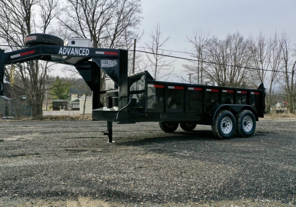A 14K Gooseneck Low Profile Dump Trailer with dual axles is parked on gravel. It has a black, heavy-duty build and features the ADVANCED label on the front, with trees and buildings visible in the background. A 14K Gooseneck Low Profile Dump Trailer with dual axles is parked on gravel. It has a black, heavy-duty build and features the ADVANCED label on the front, with trees and buildings visible in the background.