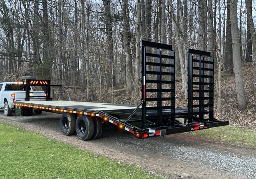 A white pickup truck is towing a large flatbed trailer with dual axles and ramp extensions, which has amber and red lights along its sides. This trailer for sale is set on a gravel path beside a grassy area, with tall Ohio trees in the background. A white pickup truck is towing a large flatbed trailer with dual axles and ramp extensions, which has amber and red lights along its sides. This trailer for sale is set on a gravel path beside a grassy area, with tall Ohio trees in the background.