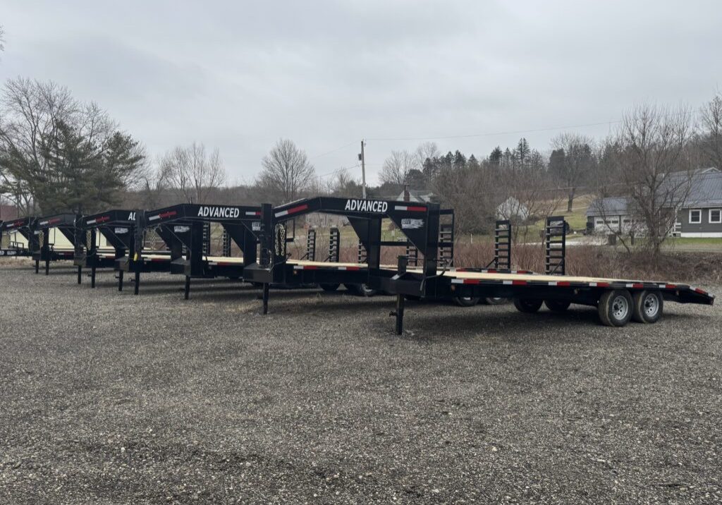 A row of three flatbed trailers, each with ADVANCED on the goosenecks and marked as trailers for sale, are parked on a gravel lot. In the background, leafless trees and a few houses sit under a cloudy sky.