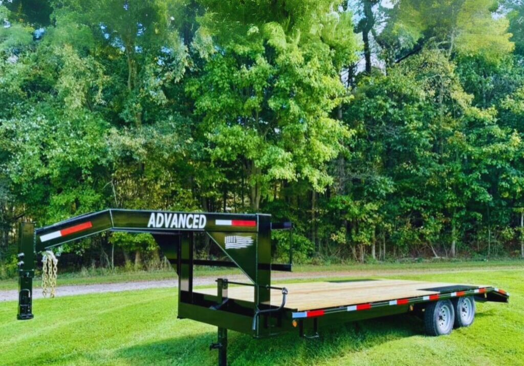 A flatbed trailer labeled Advanced is parked on a grassy area. Its surrounded by tall green trees with a clear blue sky overhead. The trailer has a black frame with a wooden deck and two sets of wheels. Different types of gooseneck flatbed trailers lined up.