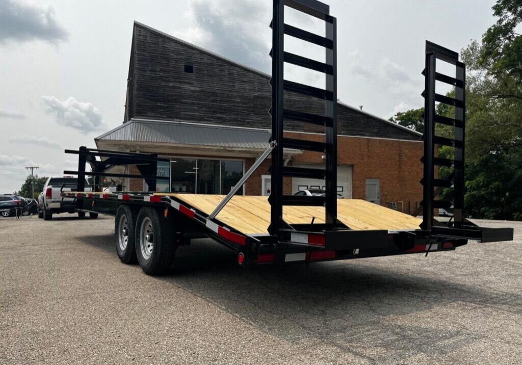 A flatbed trailer with a wooden deck and two metal ramps stands in a parking area in front of a rustic building. A red and white truck is partially visible to the left. The sky is cloudy, with some trees on the right. A comparison of gooseneck flatbed trailers with other trailer types.