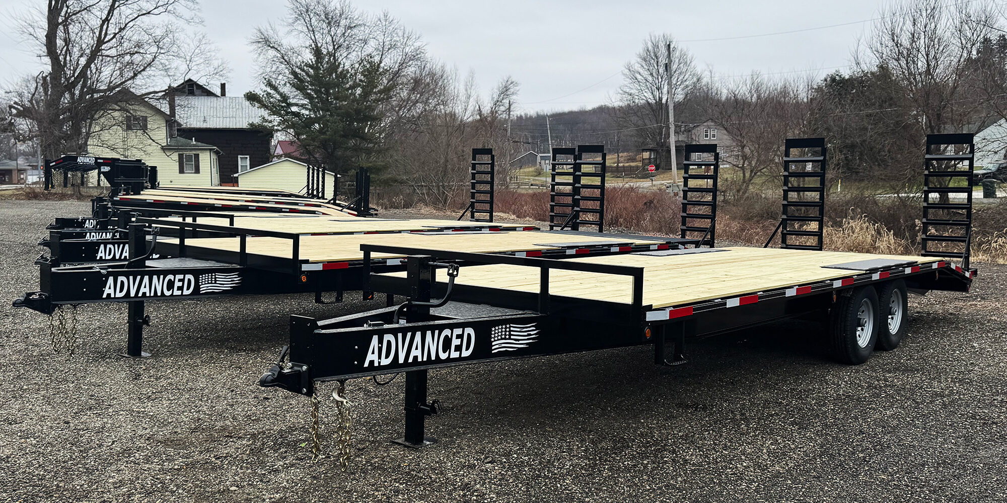 Two flatbed trailers for sale with wooden decks, labeled Advanced, are parked on a gravel lot. Each trailer features two axles and rear ramp gates. Leafless trees and a house complete the backdrop under a cloudy sky.