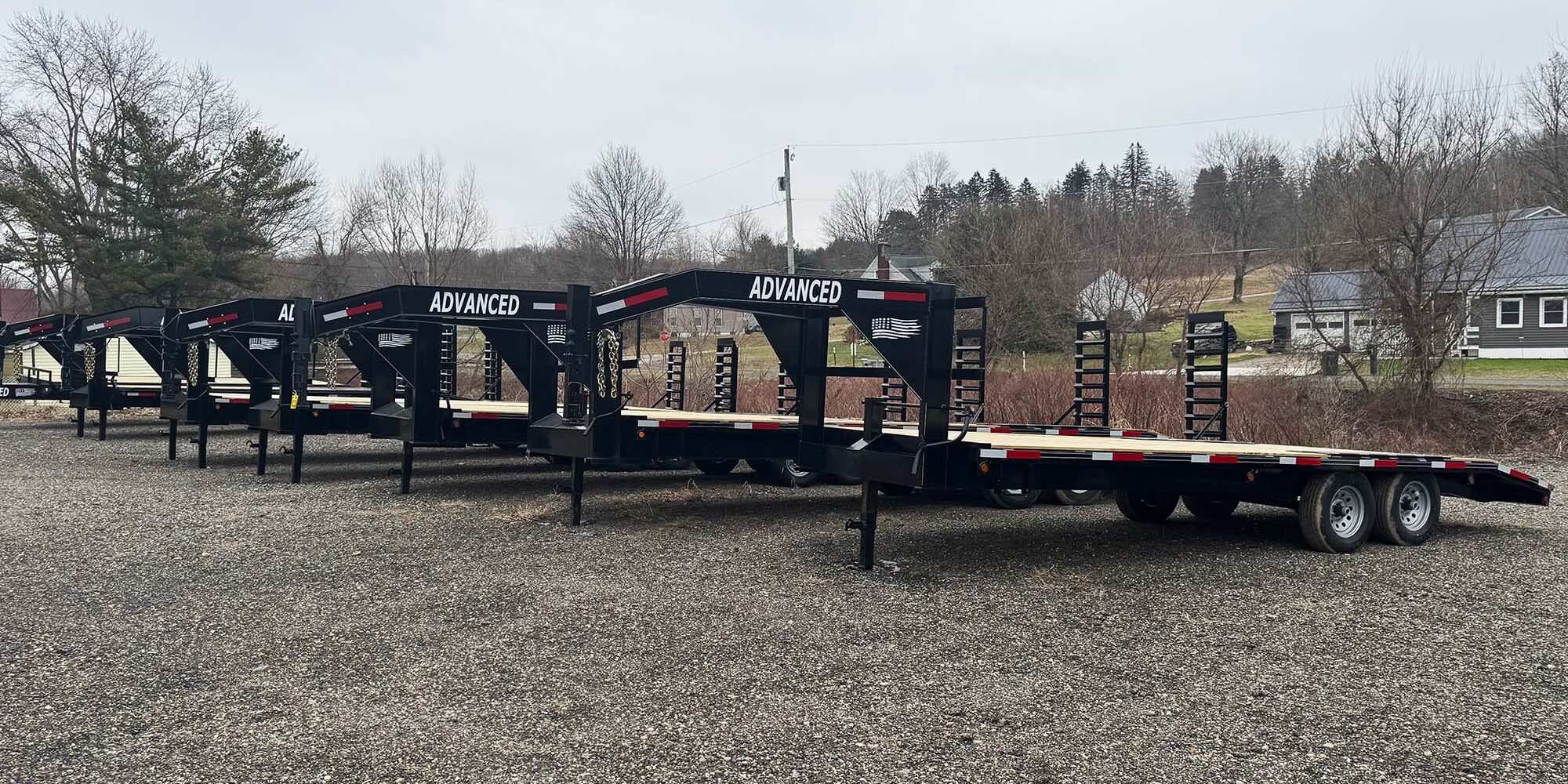 A row of black flatbed trailers labeled ADVANCED is lined up in a gravel lot, set against a backdrop of houses and trees under a cloudy sky. These trailers for sale are ready for their next journey.